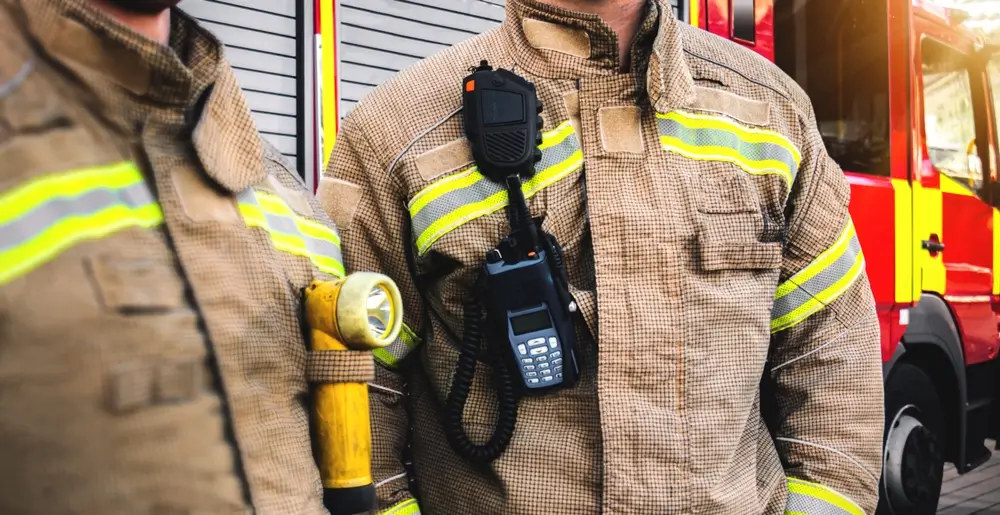 Équipe de pompiers en uniforme d'intervention avec radio et lampe torche devant leur véhicule de secours rouge.