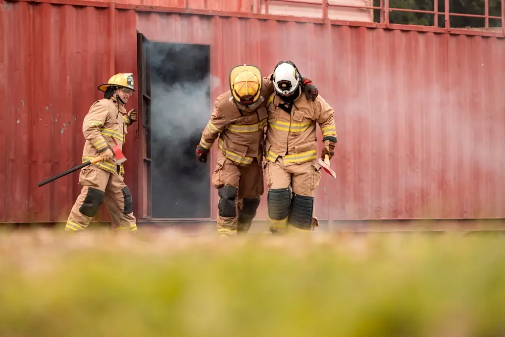 Pompiers en tenue de protection évacuant un collègue lors d'un exercice d'entraînement incendie devant un conteneur enfumé.