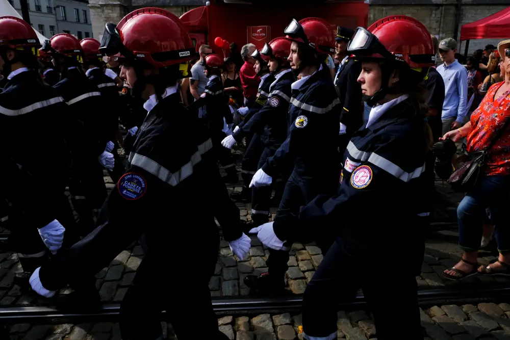 Pompiers belges défilant en uniforme avec casque rouge, illustrant la hiérarchie des grades en zone de secours.