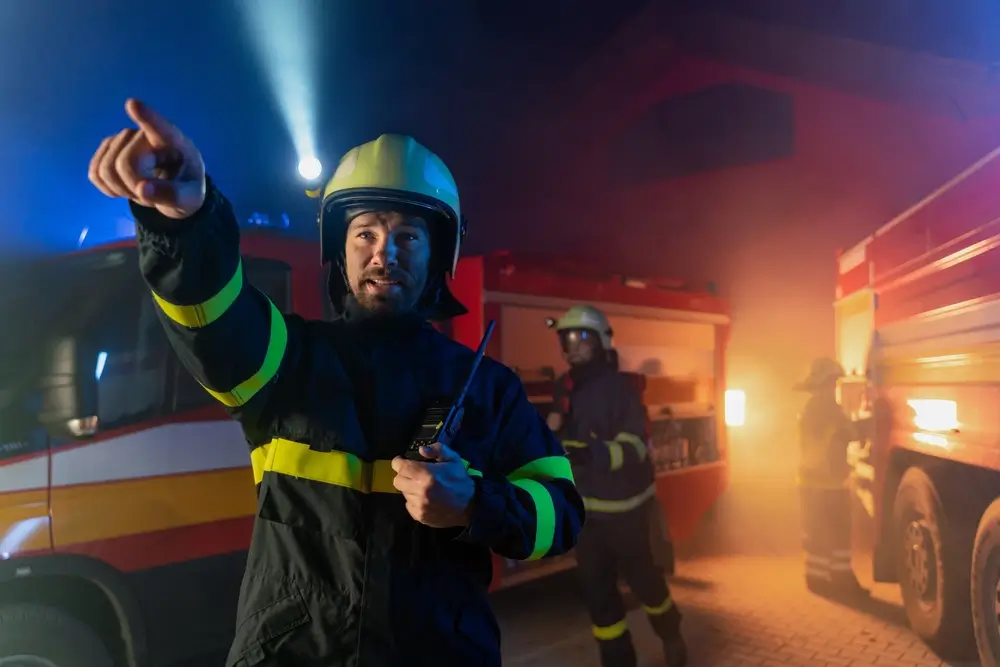 Officier pompier dirigeant une intervention avec son équipement radio devant des véhicules de secours.