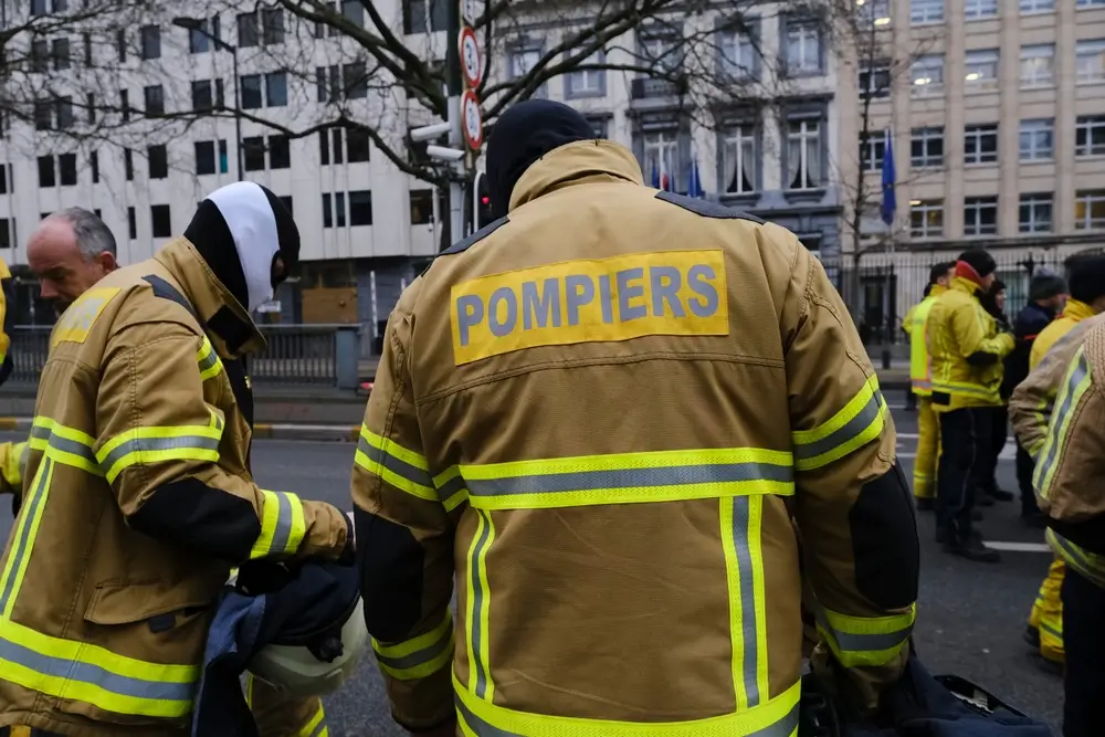 Pompiers belges vêtus de l'uniforme d'intervention beige avec bandes réfléchissantes et marquage dorsal jaune.