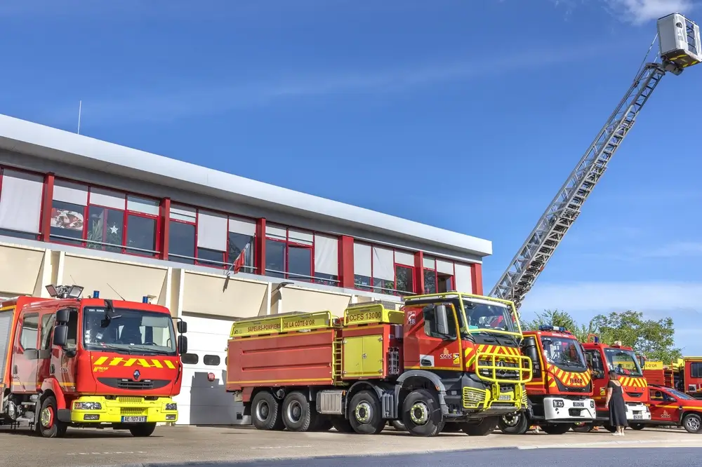 Plusieurs véhicules de secours garés devant le garage d'une caserne de pompiers avec une grande échelle déployée.